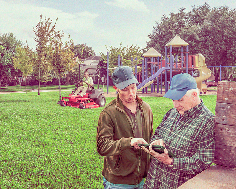 Landscaper mowing in the background with two other men look at landscaping schedule on a tablet